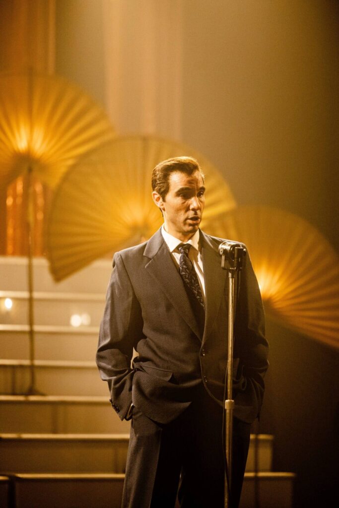 Muž v obleku stojící před zlatými dekorativními vějíři, záběr z filmu.